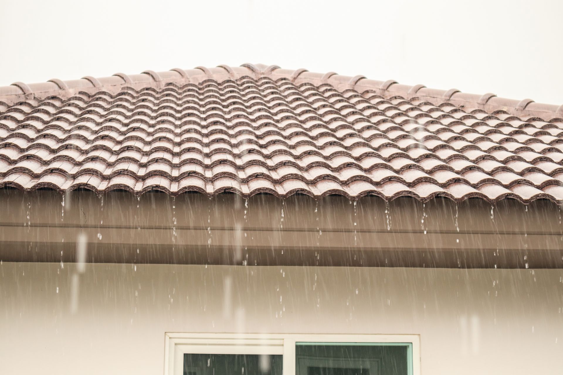 Pluie tombant sur le toit d'une maison en tuiles brunes, au-dessus d'une fenêtre