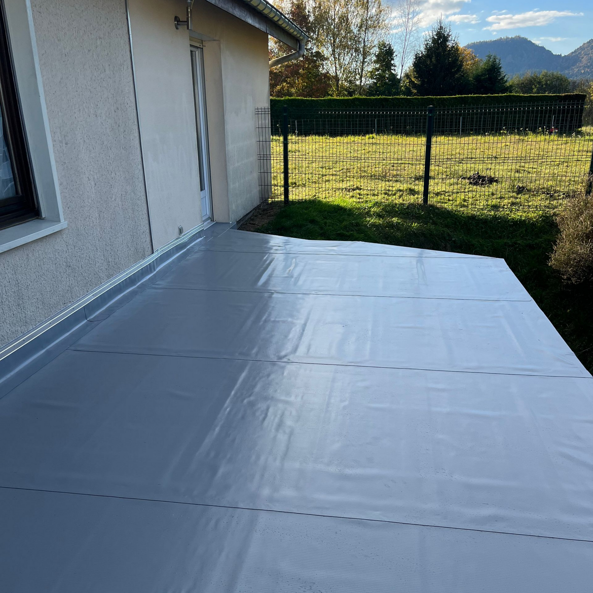 Une feuille de protection est étendue sur une terrasse à côté d'une maison, donnant sur une cour clôturée et ensoleillée avec les montagnes à l'horizon