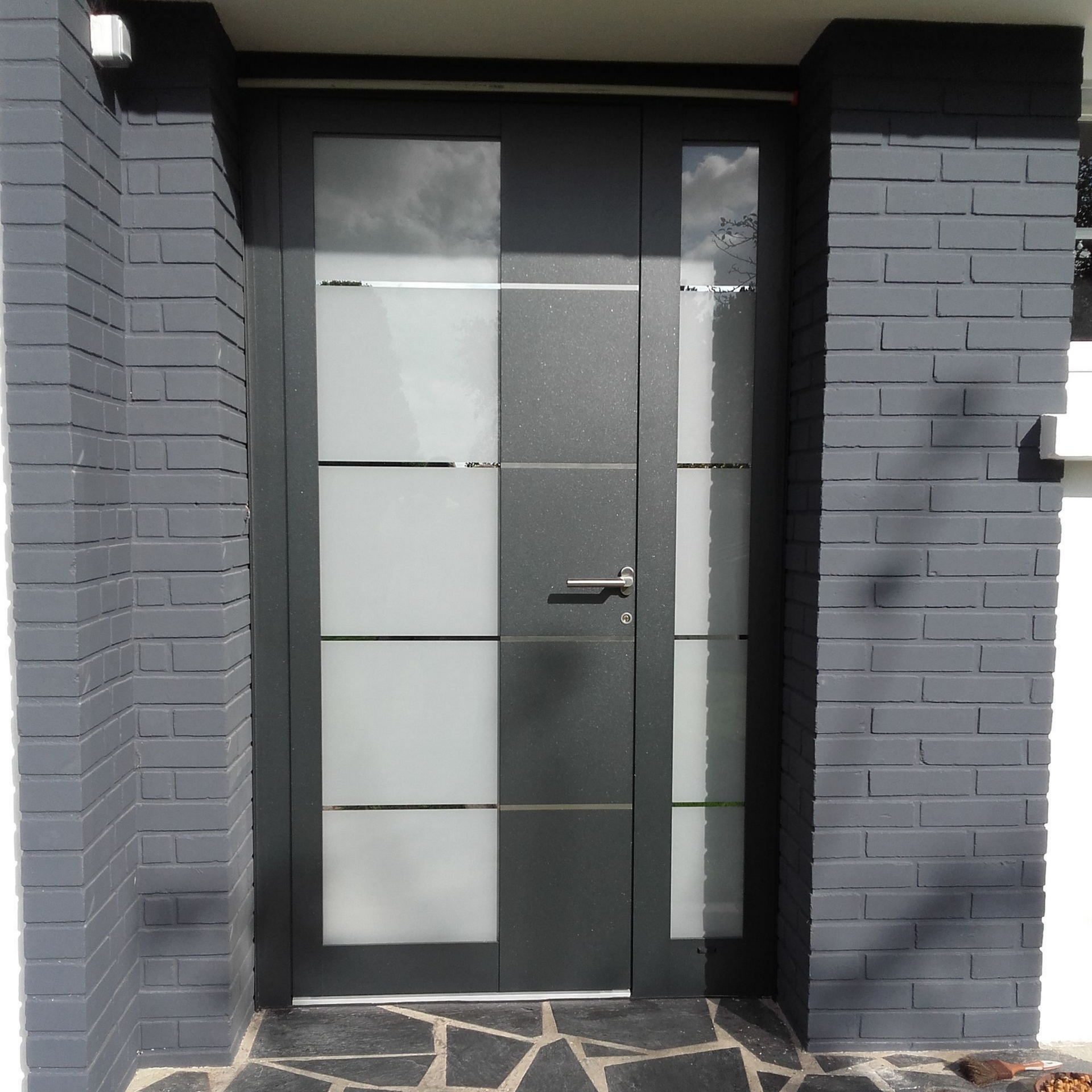 Porte d'entrée gris foncé avec panneaux de verre dépoli, flanquée de colonnes en briques grises peintes.