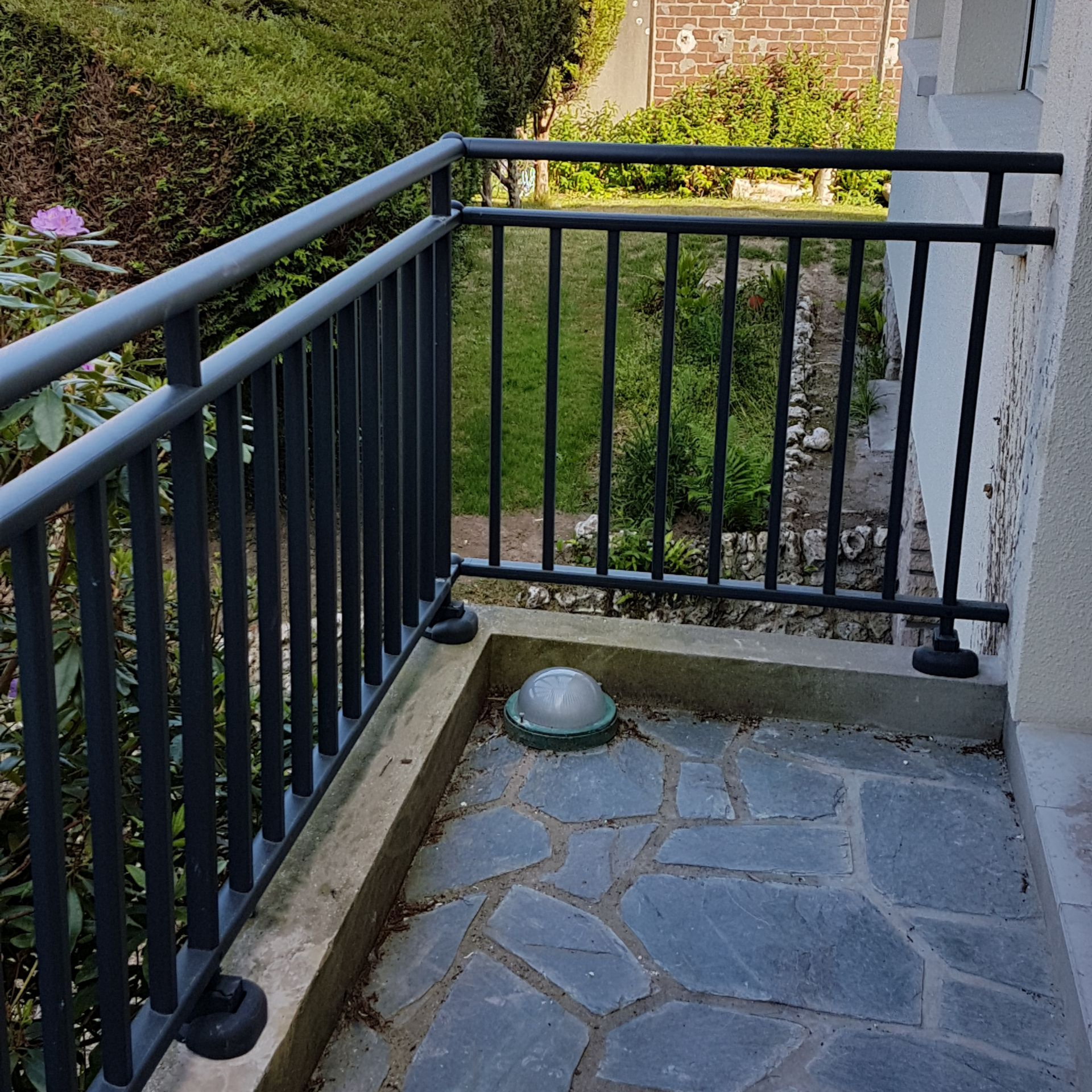 Un petit balcon avec une rambarde noire, un sol en pierre et une vue sur le jardin.