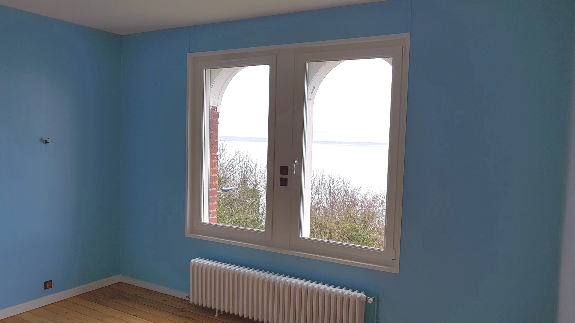 Chambre bleu clair avec une fenêtre donnant sur un plan d'eau ; un radiateur se trouve en dessous.