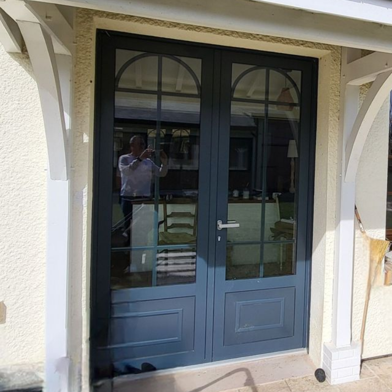 Double porte grise à panneaux de verre arqués donnant sur l'intérieur ; une personne prenant une photo se reflète dans la porte.