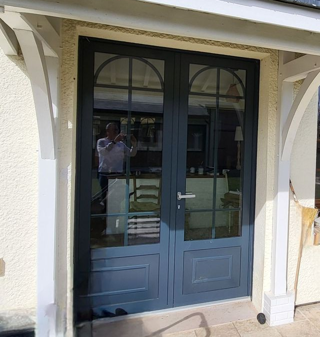 Double porte bleue à panneaux de verre cintrée, sous un auvent blanc. Une personne se reflète dans le verre.