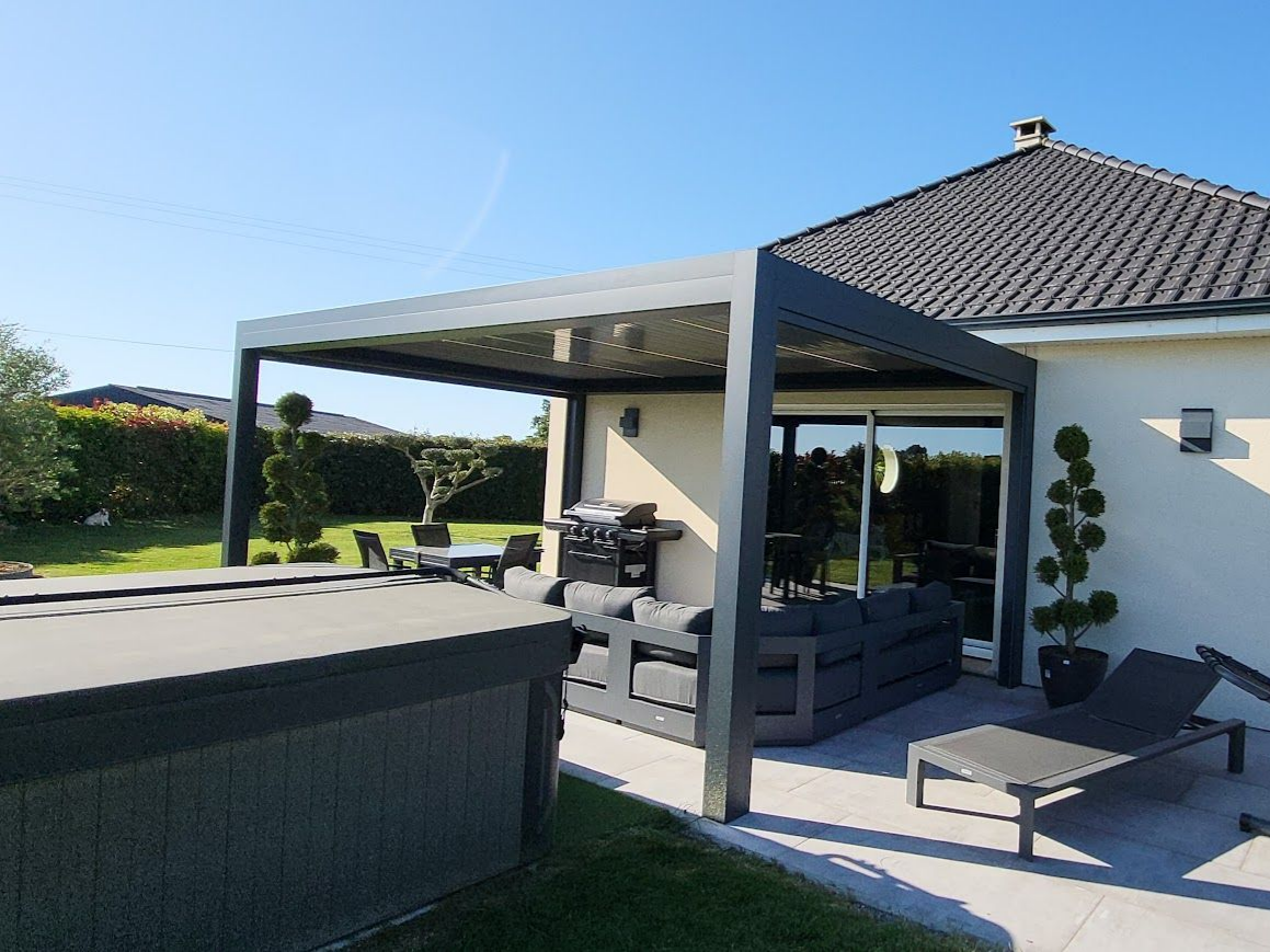 Une pergola jouxte une maison avec du mobilier de jardin sur une terrasse sous un ciel d'un bleu éclatant.