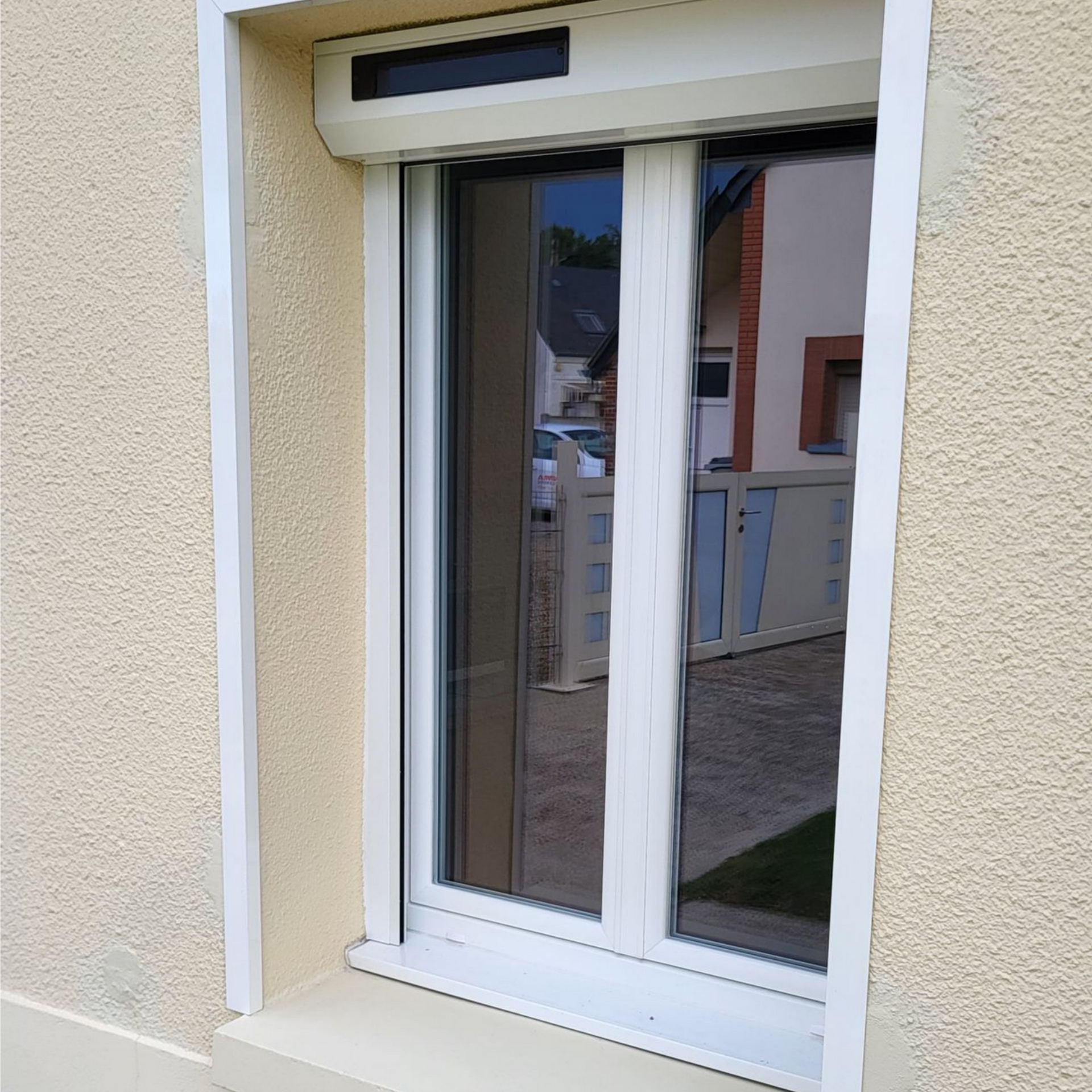 Fenêtre à cadre blanc avec stores fermés, encastrée dans une façade en stuc beige clair.