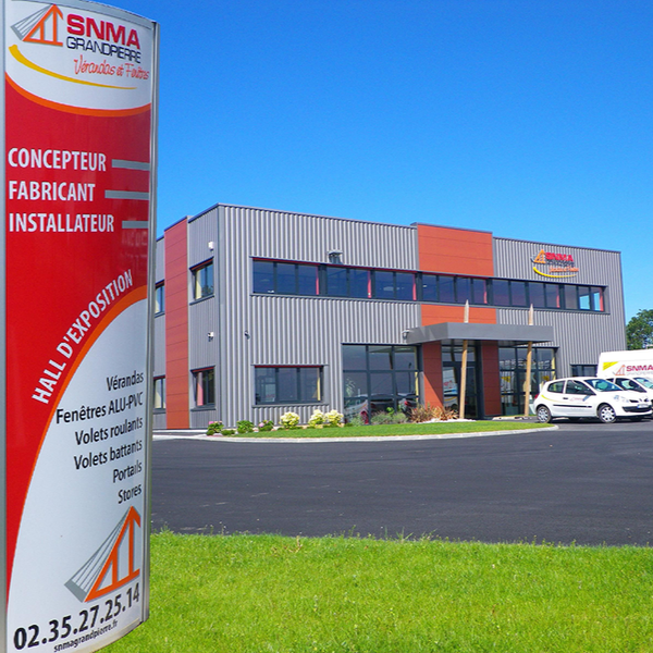 Panneau devant un bâtiment moderne, indiquant Concepteur, Fabricant, Installateur et faisant la publicité de vitrines et d'autres services.