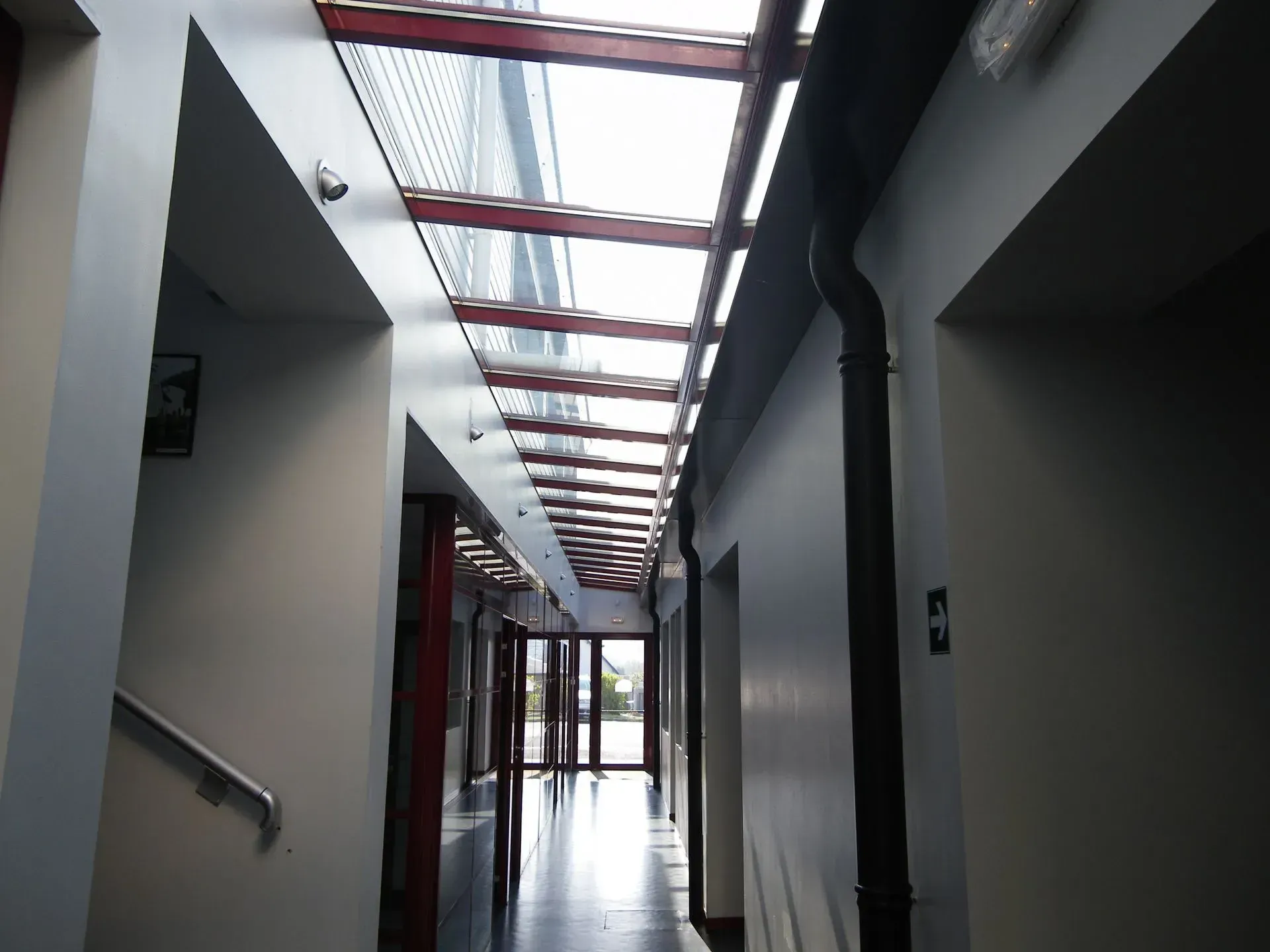 Couloir avec puits de lumière ; murs blancs, sol foncé, poutres structurelles rouges et une porte au fond.