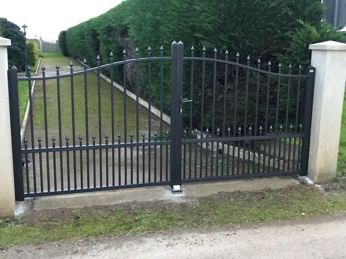 Un portail en fer forgé noir, encadré par deux piliers blancs, donne accès à une allée carrossable, avec des haies vertes en arrière-plan.