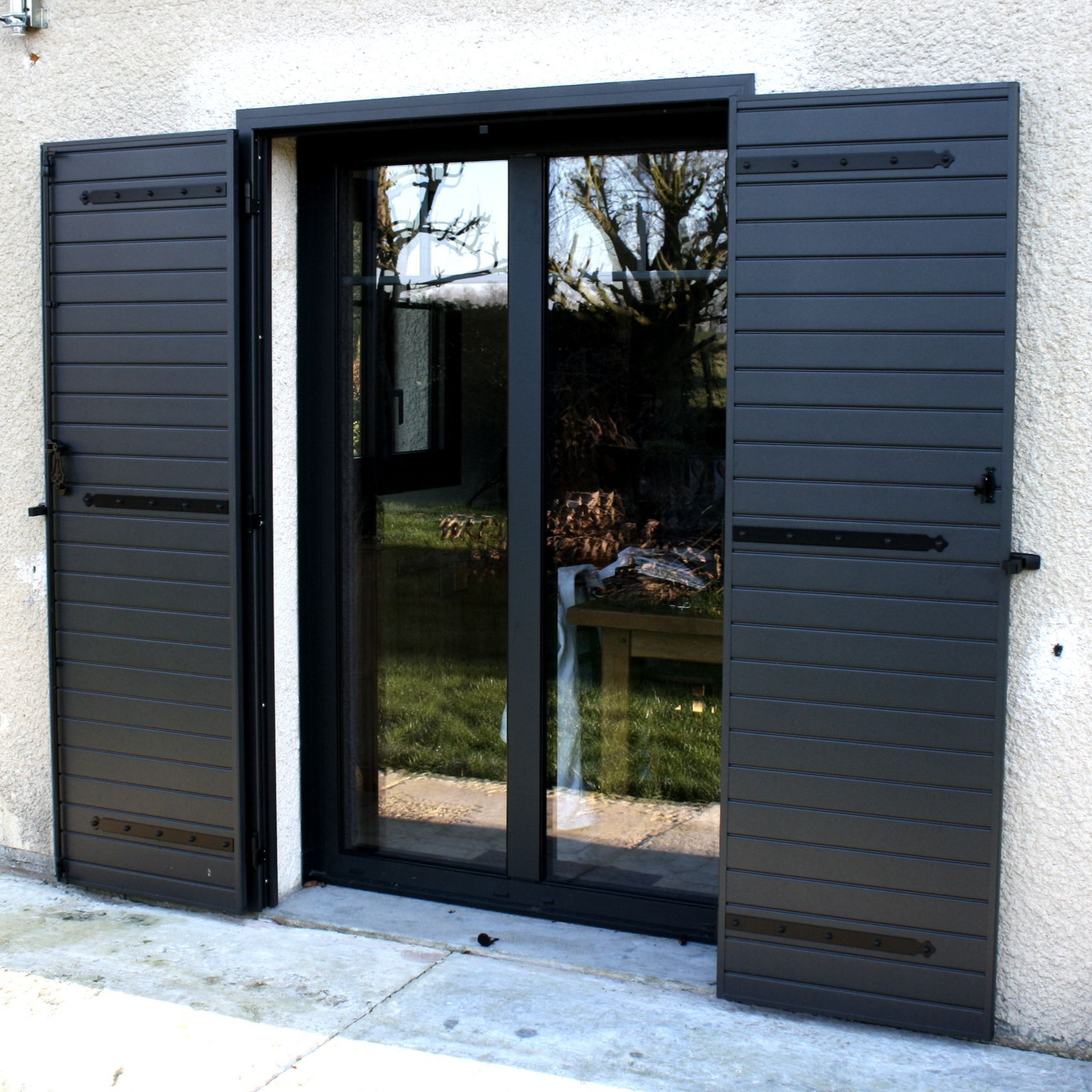 Des volets gris foncé s'ouvrent de part et d'autre d'une fenêtre à cadre noir. Ces volets sont fixés à un mur en stuc clair.