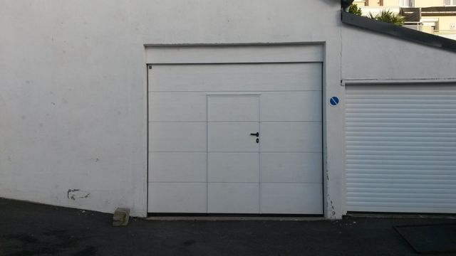 Porte de garage blanche avec une porte d'accès plus petite. Allée asphaltée et murs blancs.