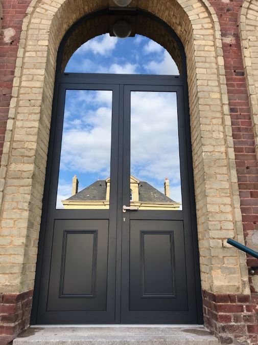 Double porte noire à arc en plein cintre, reflétant le ciel bleu et le toit de l'immeuble, intégrée à un bâtiment en briques.