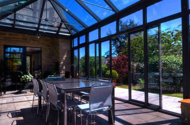 Véranda avec murs et plafond en verre, table à manger, chaises, vue sur les arbres et le ciel.