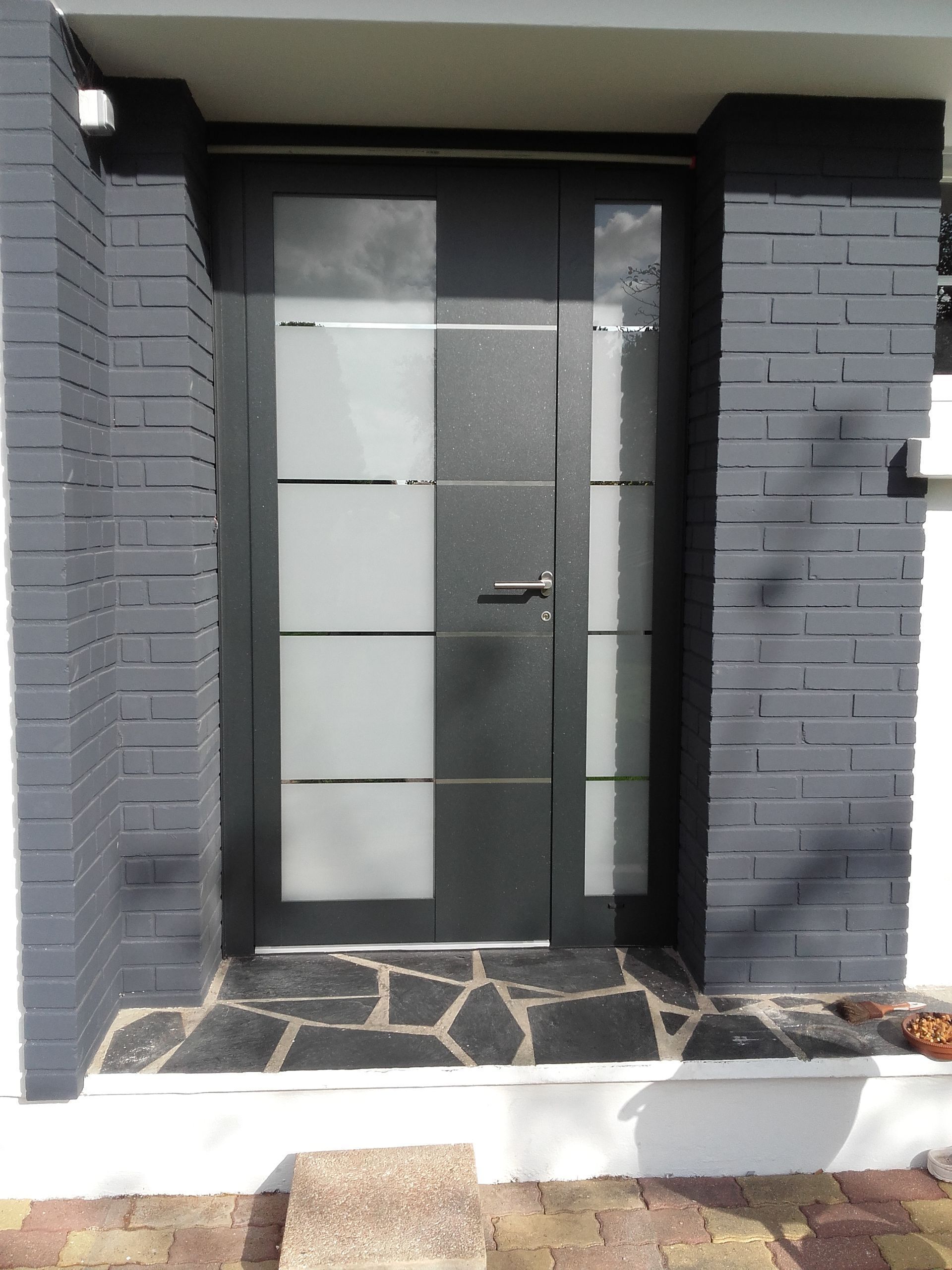Porte d'entrée noire avec panneaux de verre dépoli et allée en briques gris foncé.