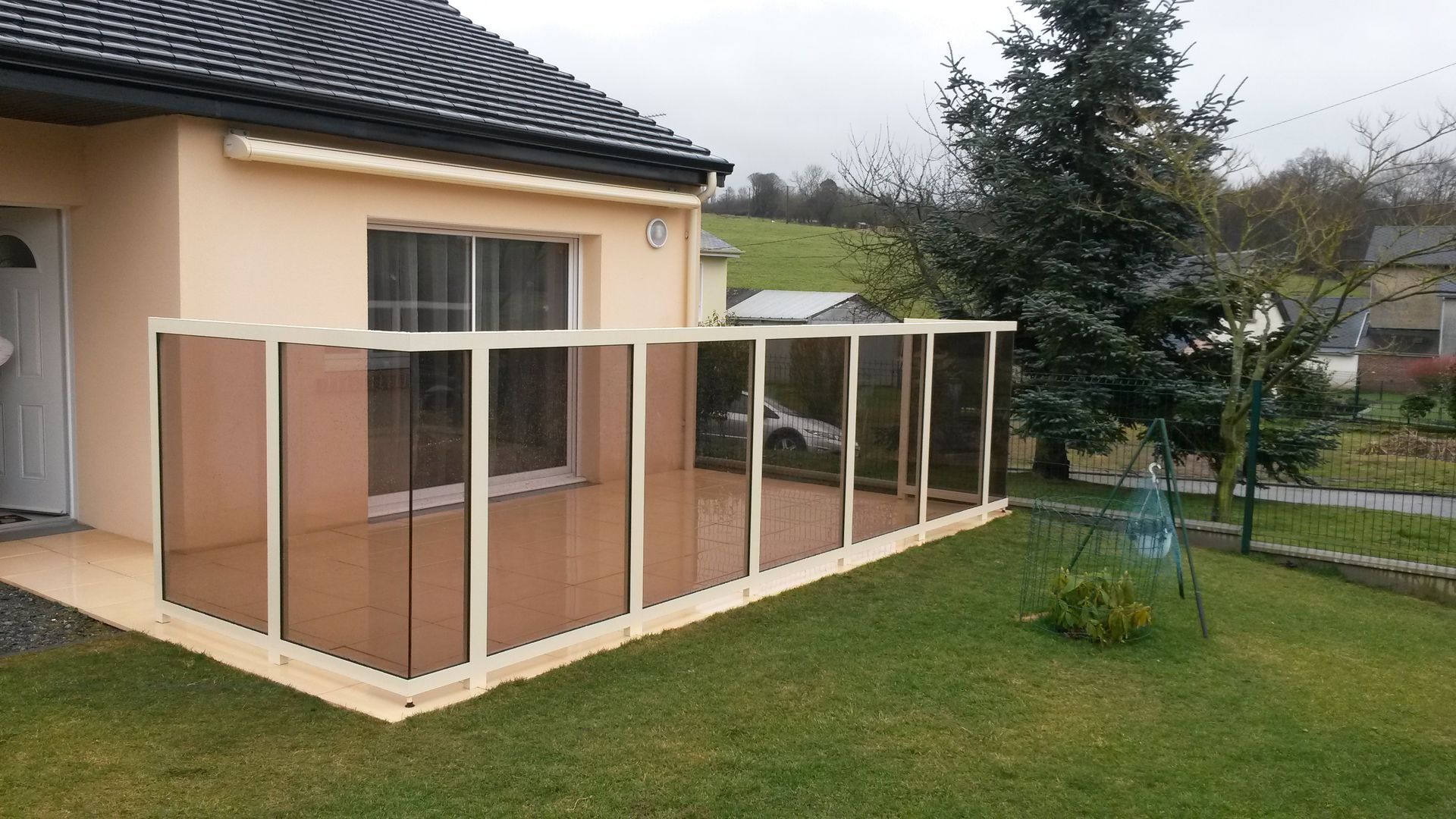 Un patio clos de panneaux teintés beiges, attenant à une maison beige, entouré de pelouse verte.