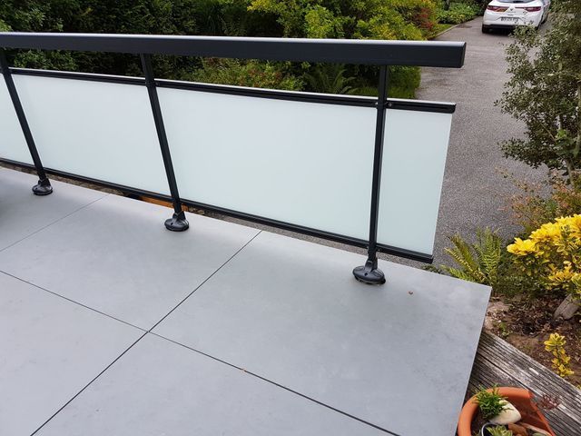 Balcon avec panneaux de verre dépoli, rambarde noire et sol carrelé gris ; voiture en arrière-plan.