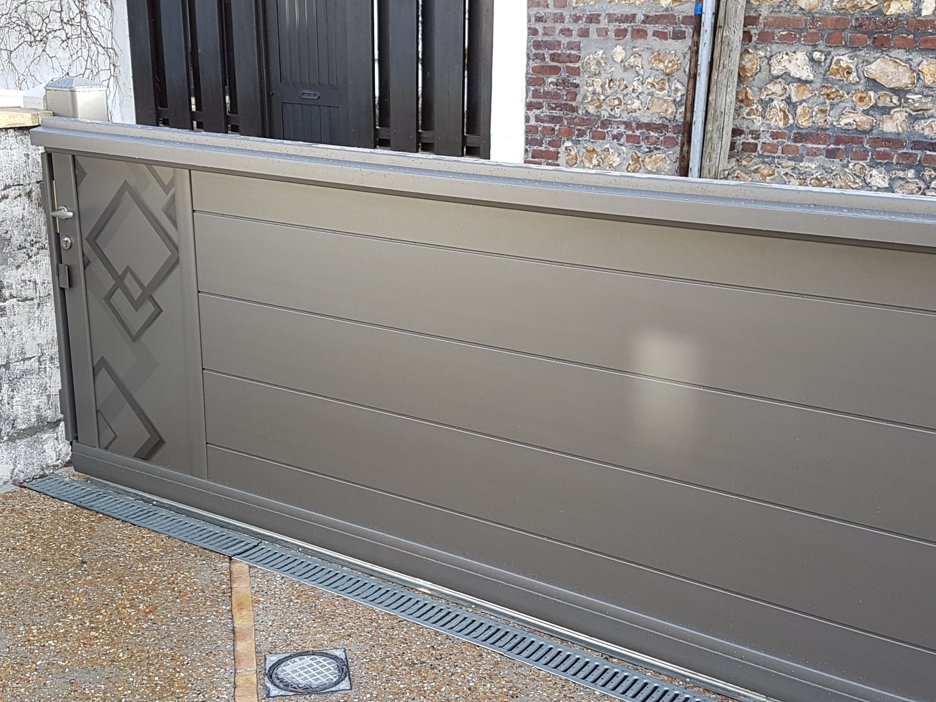 Portail coulissant en métal gris orné de motifs en losange, installé devant un mur en pierre.