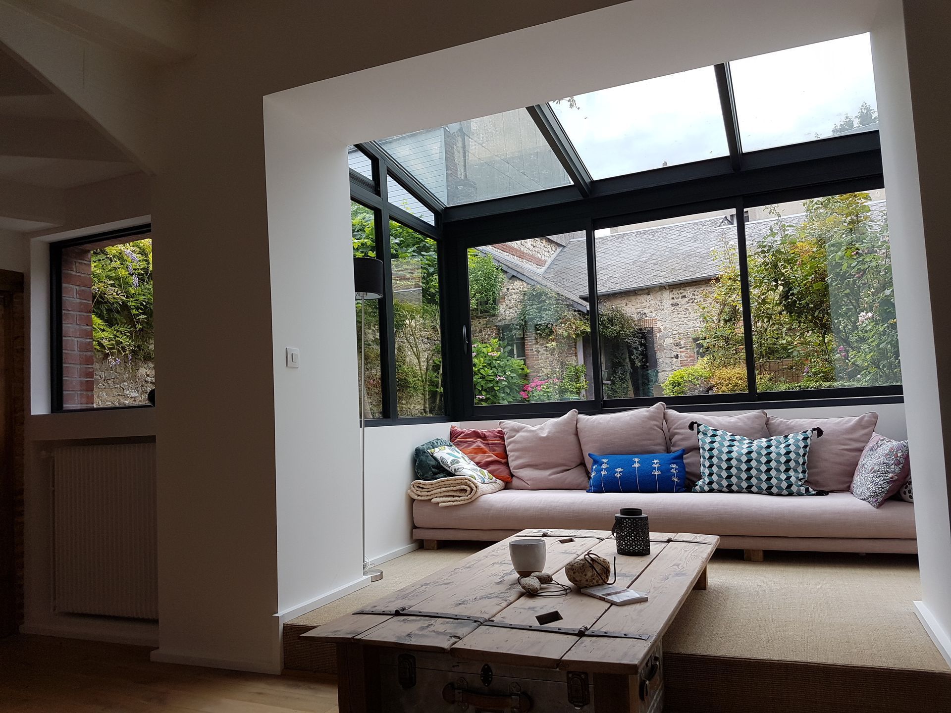 Véranda avec canapé rose, table en bois, toit en verre, donnant sur la verdure.