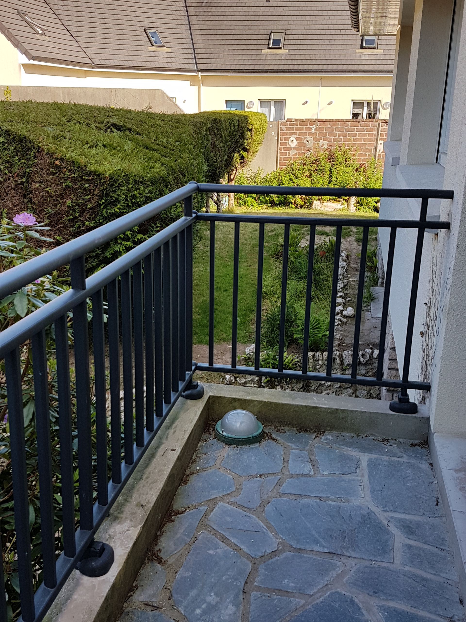 Petit balcon avec rambarde en métal noir, sol en pierre et vue sur la verdure.