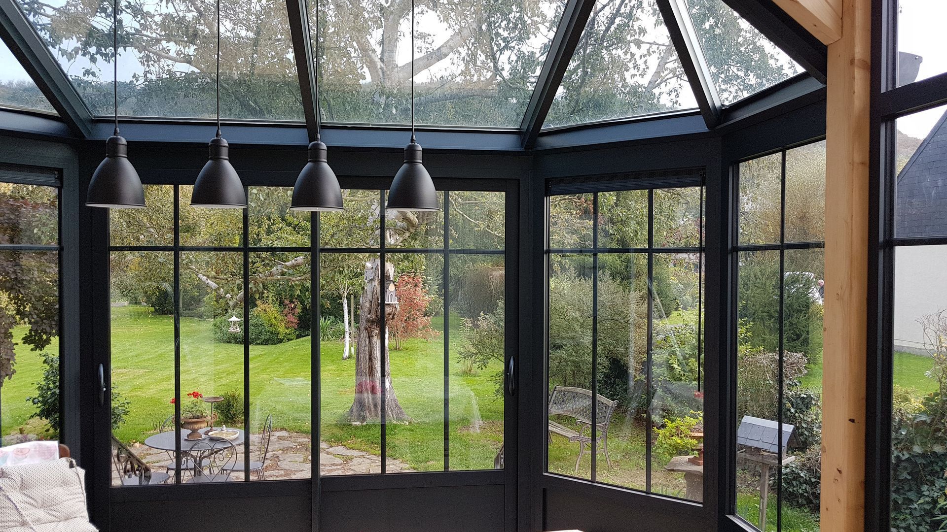 Véranda avec fenêtres à cadre noir et vue sur un jardin verdoyant. Quatre suspensions noires sont installées à l'intérieur.