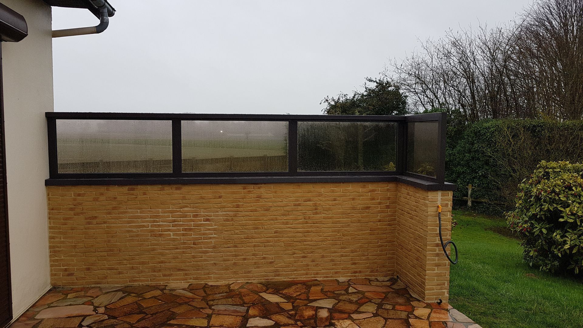 Patio avec mur de briques, balustrade en verre et ciel couvert.