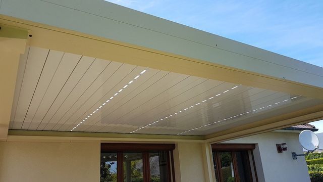 Toiture de patio beige à lames horizontales, attenante à un bâtiment clair. Ciel bleu visible.