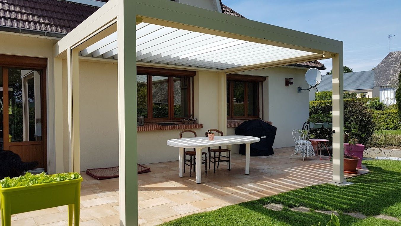 Pergola beige au-dessus d'un patio avec une table et des chaises, attenante à une maison avec une pelouse verte.