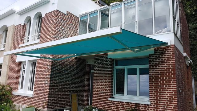 Un auvent turquoise s'étend depuis l'entrée d'un bâtiment en briques. On aperçoit des fenêtres claires.