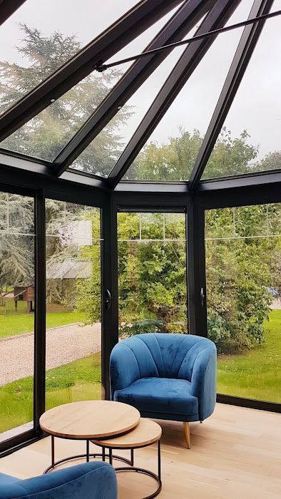 Véranda avec chaises bleues, petites tables et vue sur des arbres verts à travers les baies vitrées.