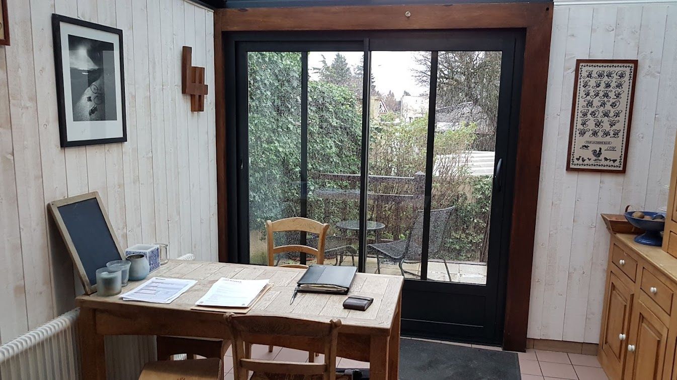 Véranda avec table en bois, chaises et porte-fenêtre coulissante donnant sur la verdure.