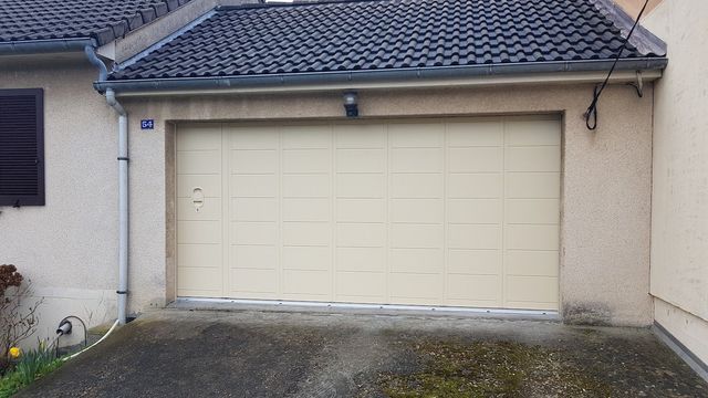Garage avec une porte beige, sur un bâtiment beige clair sous un toit foncé.