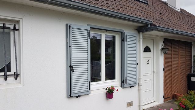 Maison blanche avec volets et porte gris, petit pot de fleurs sous la fenêtre.