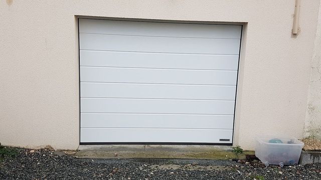 Porte de garage blanche à lames horizontales encastrée dans un mur beige. Un petit conteneur se trouve à droite.