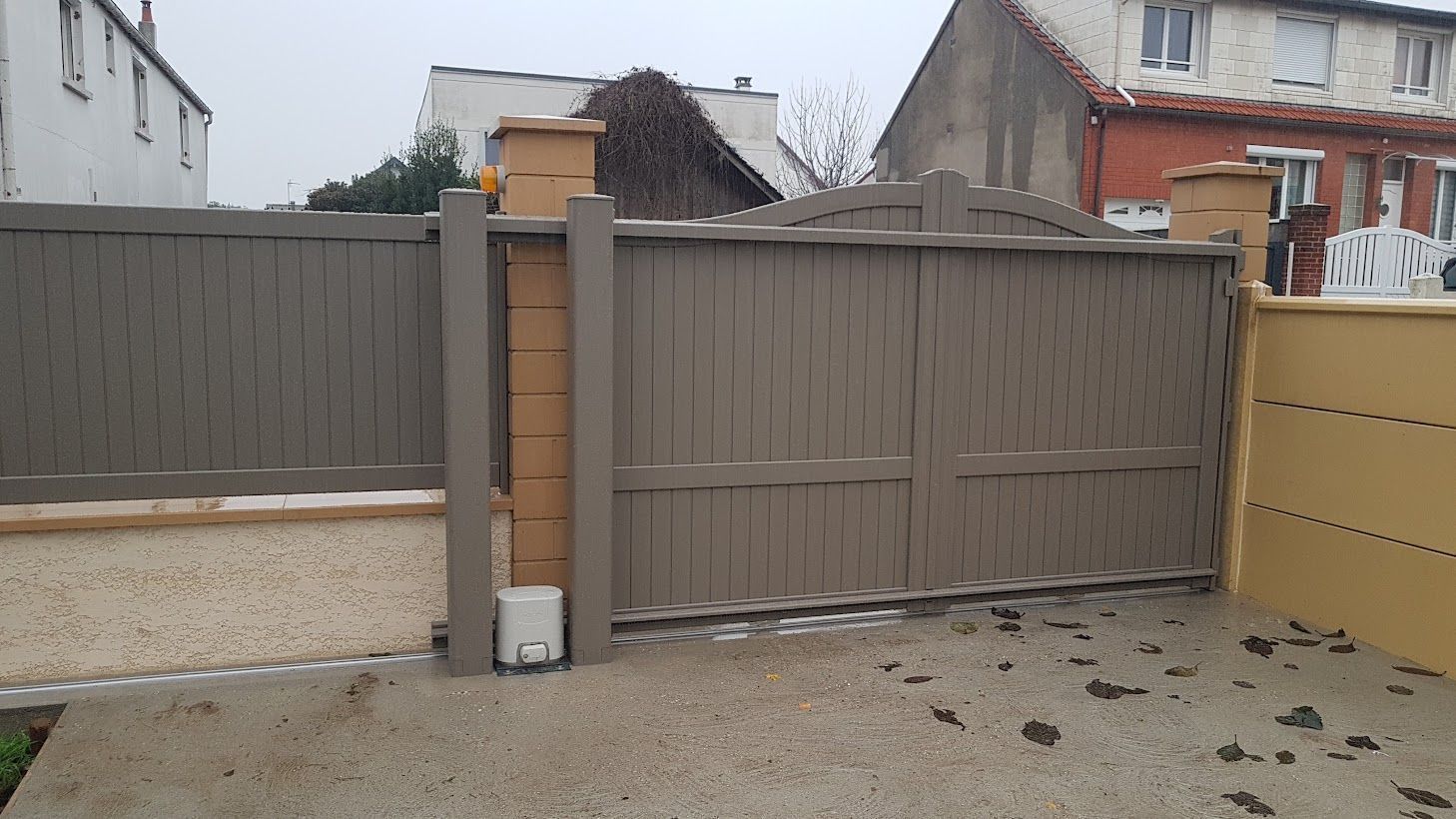 Portail d'allée en bois gris assorti à la clôture, piliers beiges et mur jaune.