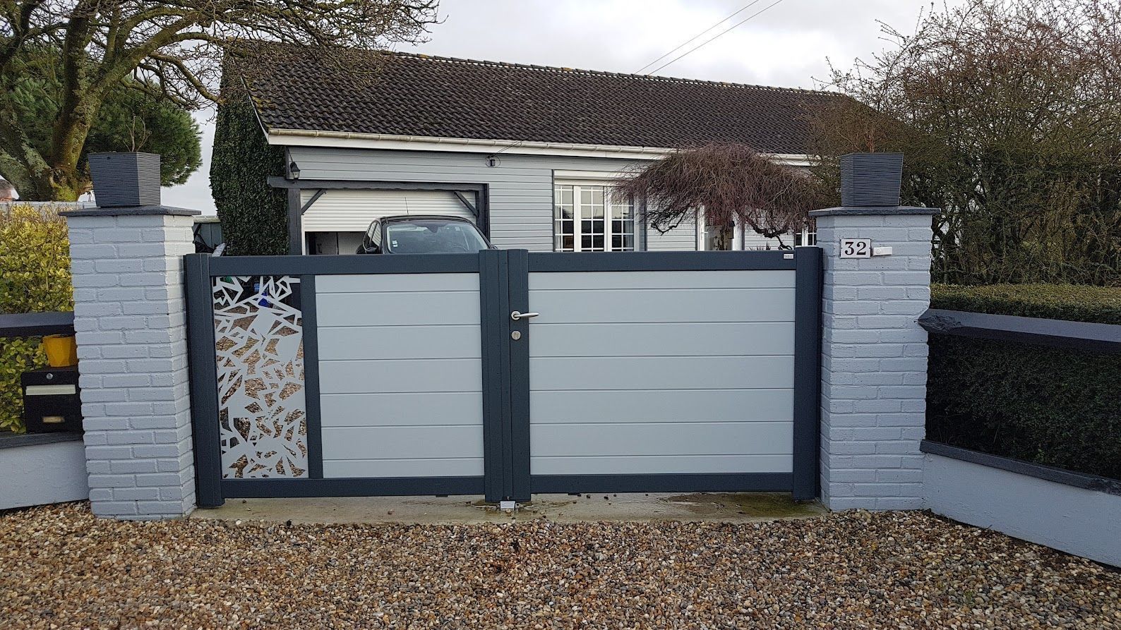 Portail et clôture en métal gris devant une maison, installés sur du gravier, avec des piliers en briques gris clair.