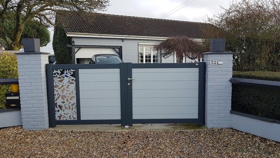Portail d'allée gris avec panneau décoratif, piliers gris et maison en arrière-plan.