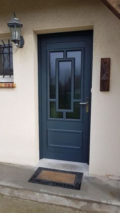 Porte d'entrée bleue avec panneau de verre, paillasson et luminaire sur un mur beige.