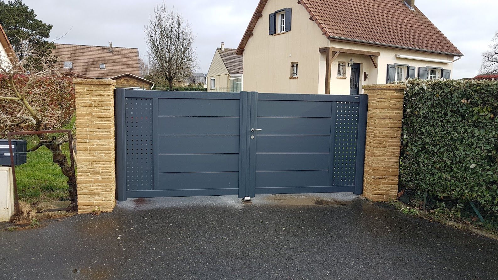 Double portail bleu foncé avec panneaux latéraux décoratifs, piliers en pierre et une maison en arrière-plan.