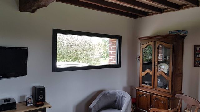 Vue intérieure d'une pièce avec une fenêtre, une télévision, une armoire en bois et un fauteuil gris.