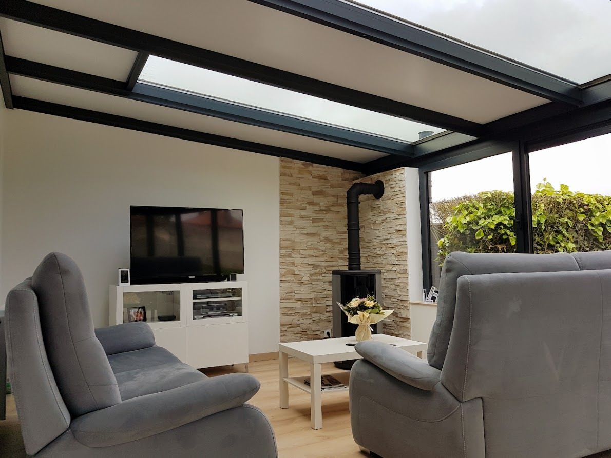 Intérieur d'une véranda avec mobilier gris, un mur en pierre et un toit en verre à cadre noir.