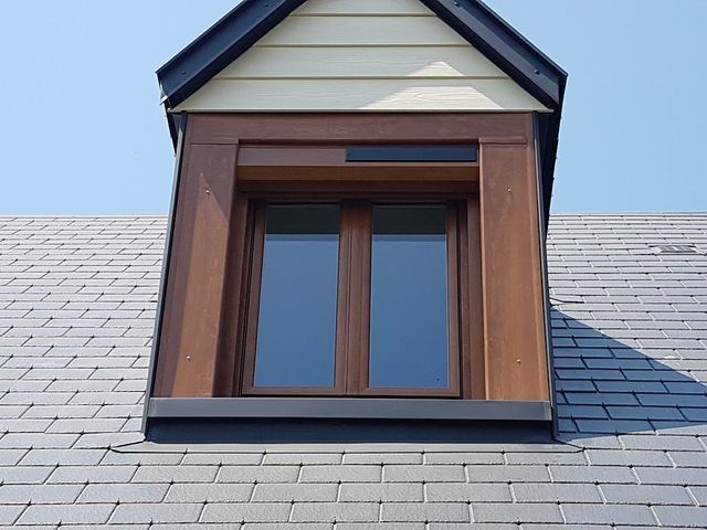 Lucarne sur un toit en ardoise grise. Un encadrement en bois brun encadre la fenêtre, et un bardage beige recouvre la toiture.