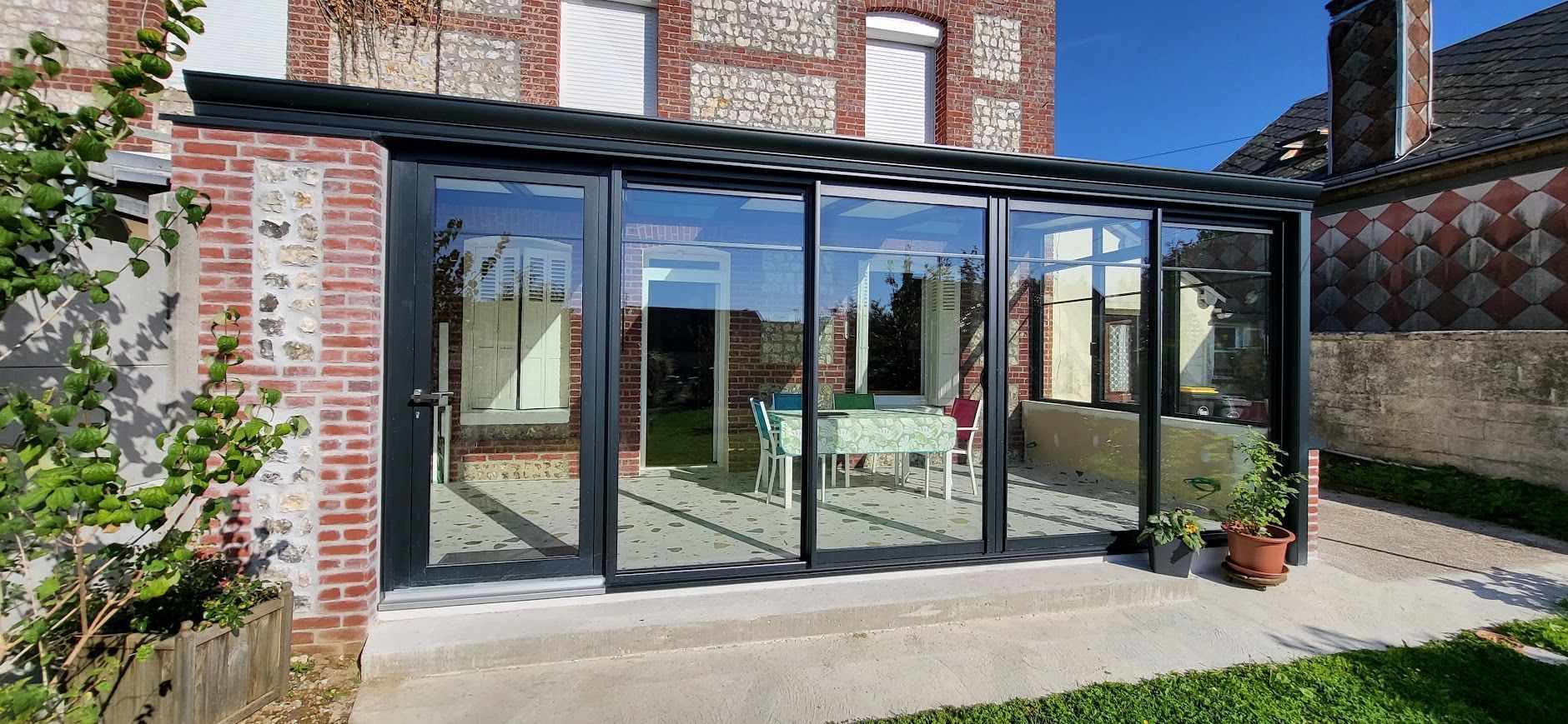 Véranda vitrée attenante à un bâtiment en briques, reflétant l'extérieur.