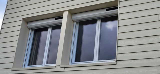 Deux fenêtres à volets roulants clairs fermés sur un bâtiment au revêtement extérieur clair.