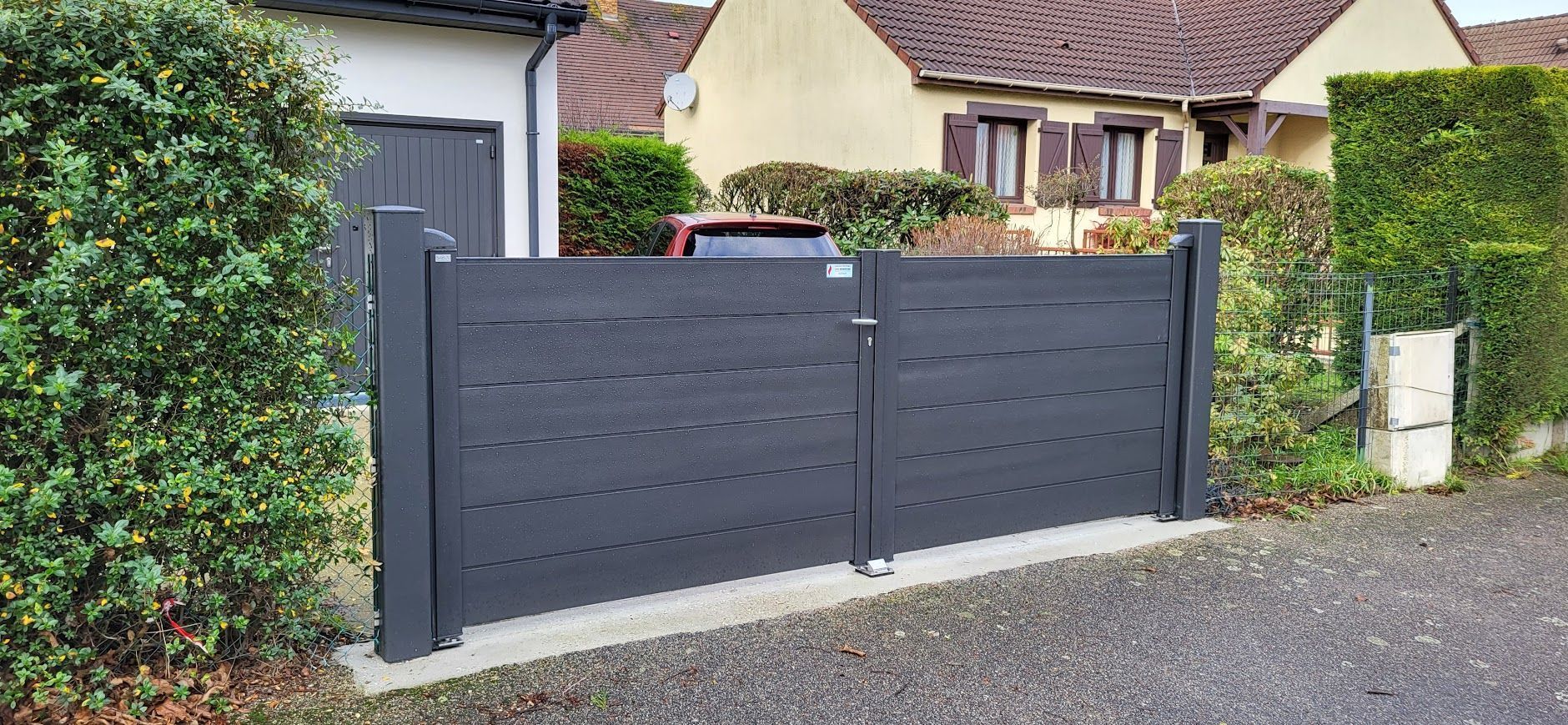 Un portail gris devant une maison, entourée de verdure.