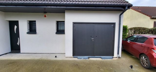 Extérieur d'une maison blanche avec une porte de garage gris foncé, une porte noire et une voiture rouge garée à proximité.