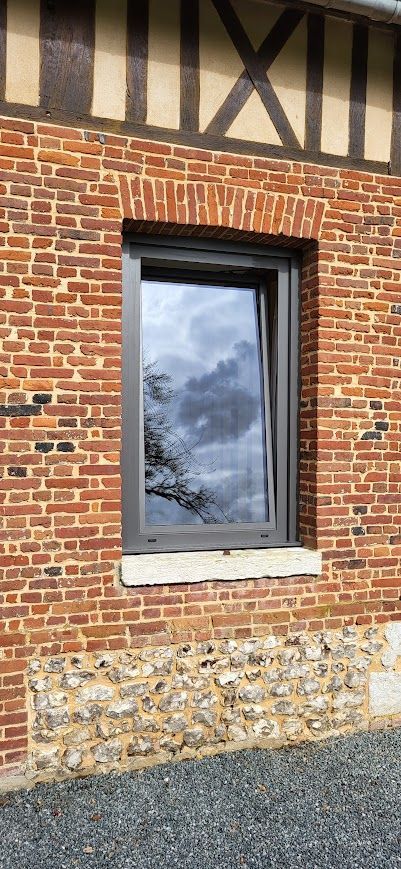 Bâtiment en briques avec une fenêtre reflétant le ciel et les arbres. Charpente en bois au-dessus de la fenêtre. Chemin de gravier en contrebas.