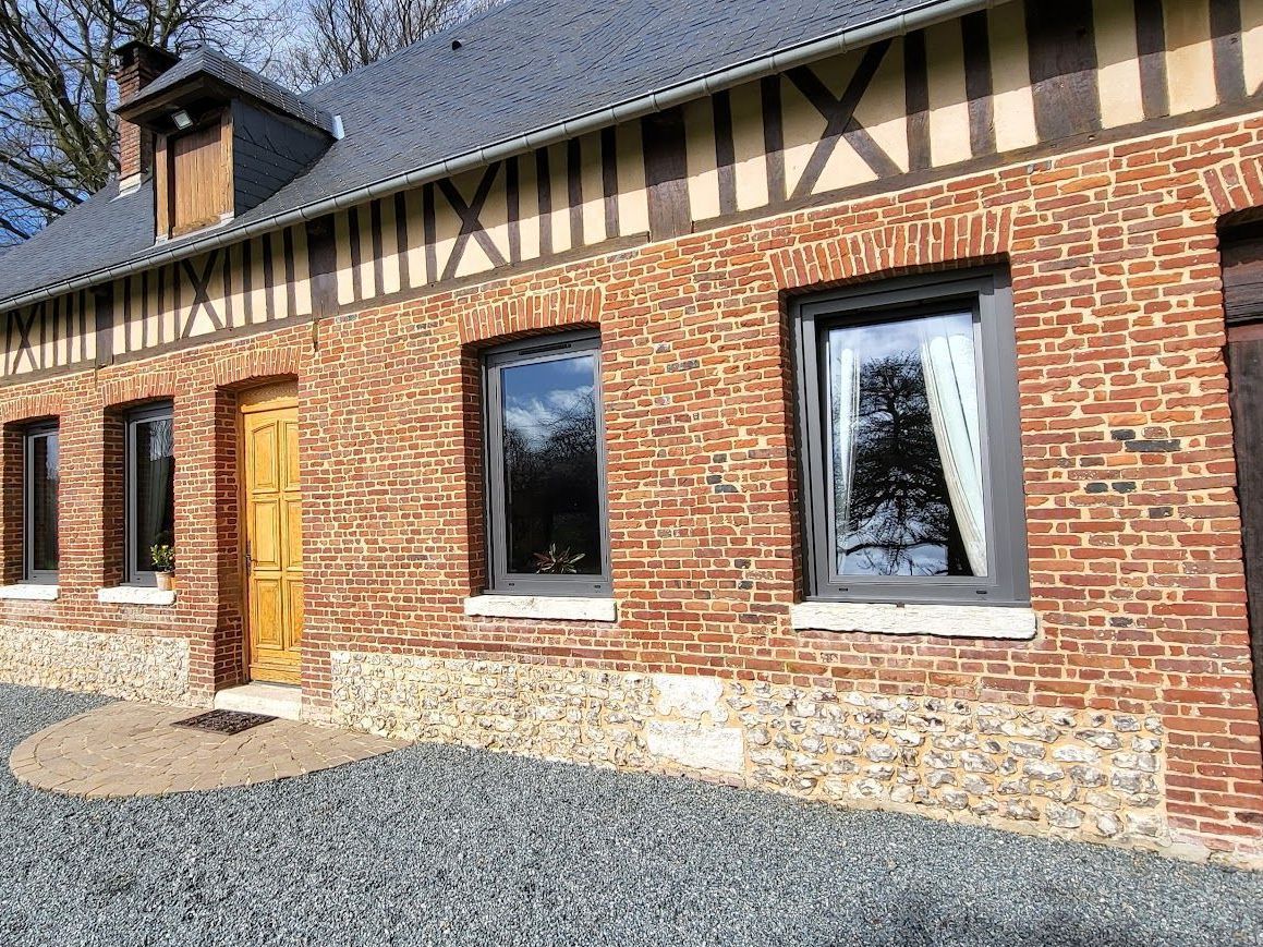 Bâtiment en briques avec charpente en bois, toit gris et fenêtres laissant apparaître le reflet des arbres.