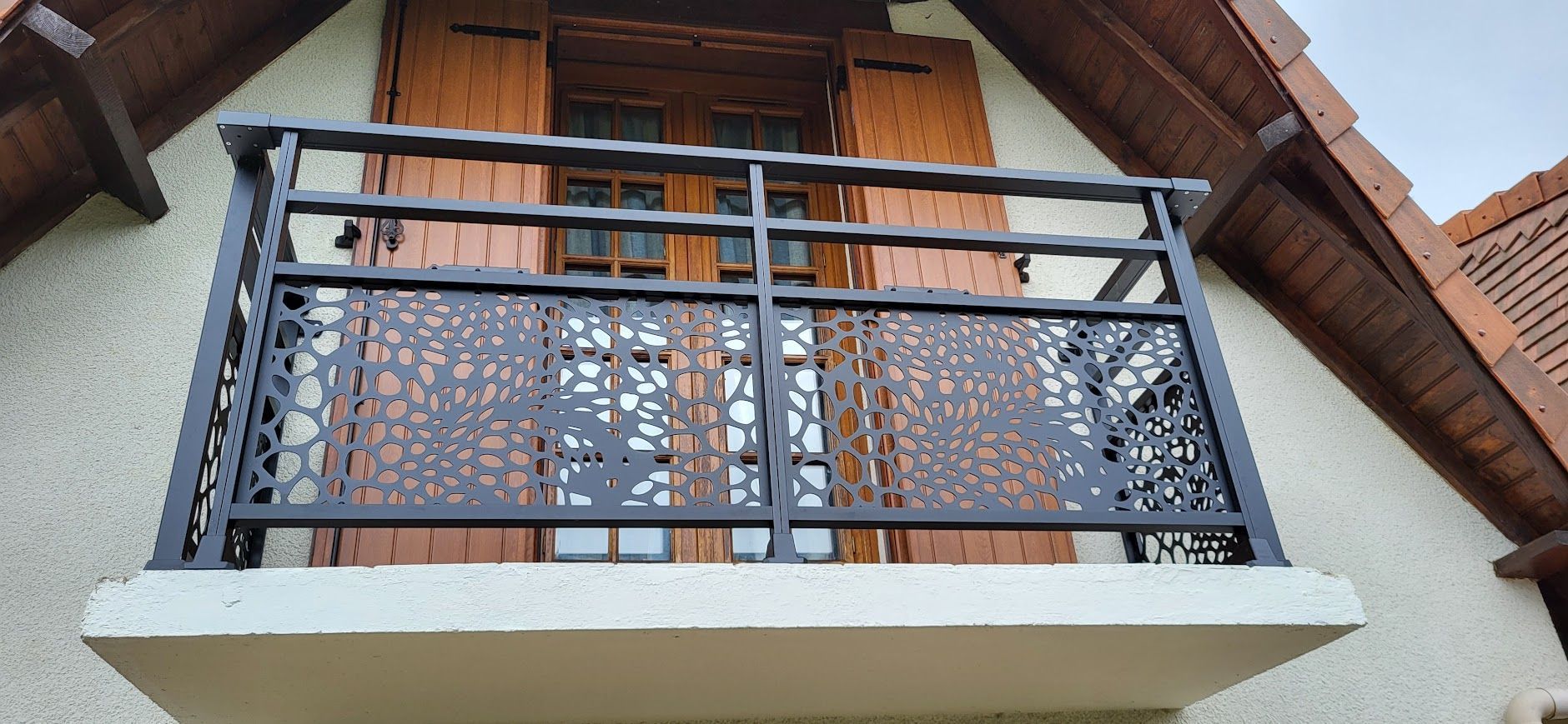 Balcon en métal noir orné de découpes décoratives sur une maison à volets en bois.