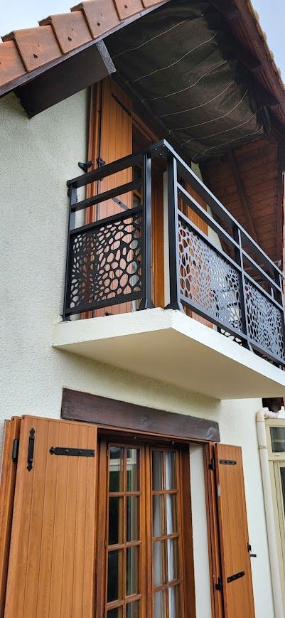 Balcon avec rambarde en métal noir et panneau décoratif sur un immeuble couleur crème avec volets en bois et toit marron.
