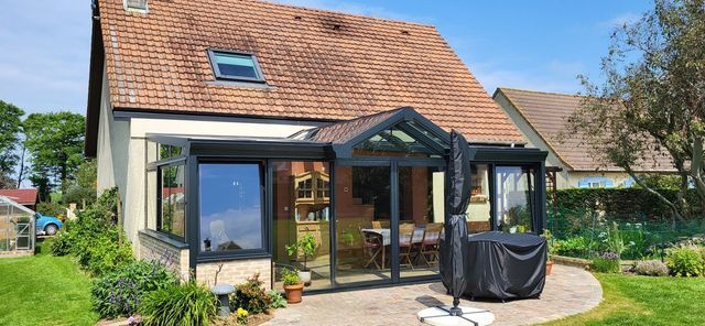 Une maison avec un patio vitré, un toit de tuiles rouges et une pelouse par une journée ensoleillée.