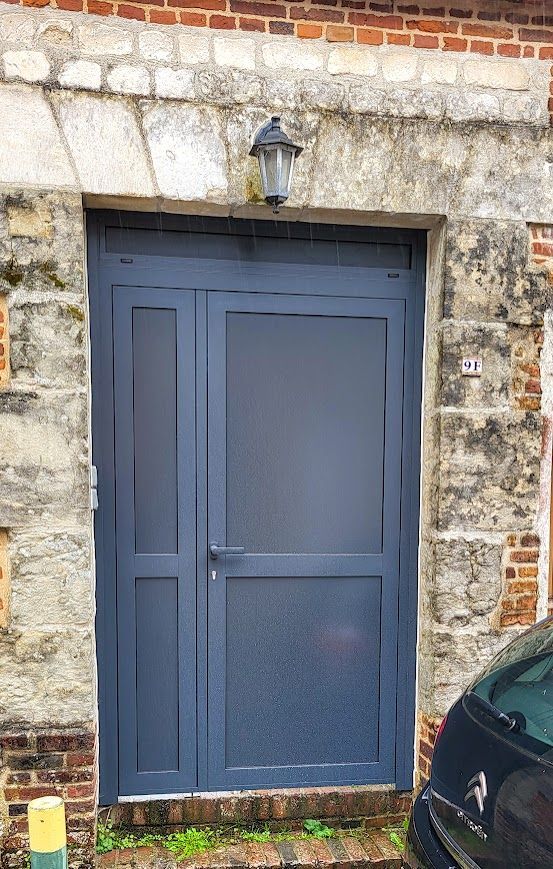 Porte d'entrée bleue avec imposte dans un bâtiment en briques et en pierres. Une voiture noire est partiellement visible sur la droite.